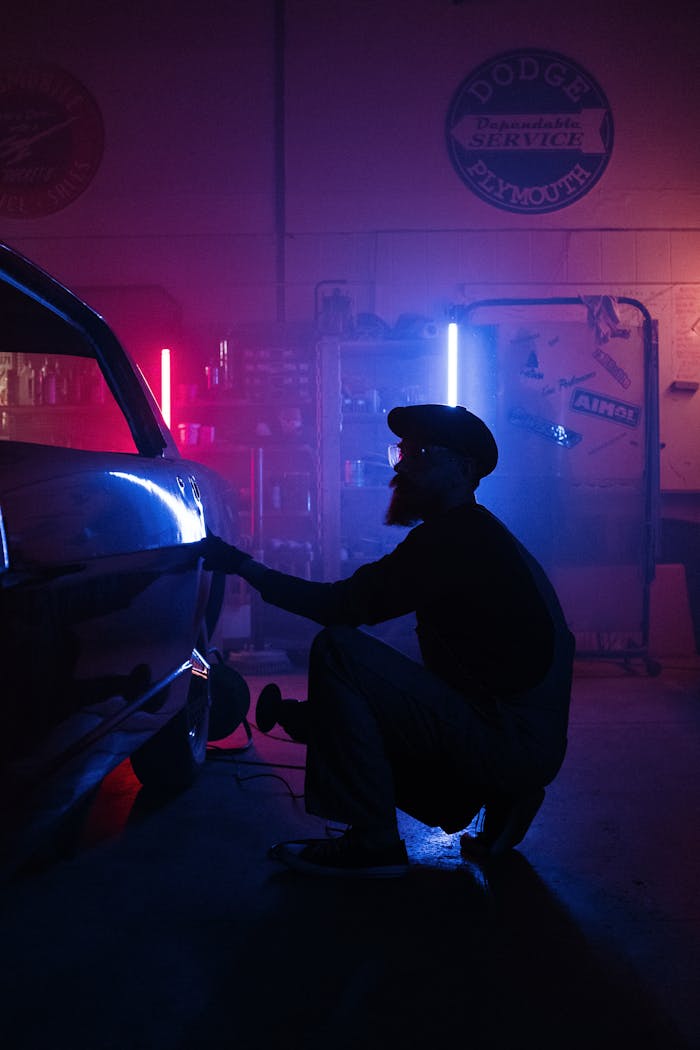 About Silhouette of mechanic repairing a car in a dimly lit garage with colorful neon lights.